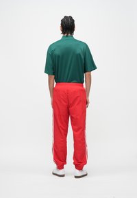 Groen overhemd met korte mouwen en kraag, gecombineerd met rode trainingsbroeken met witte zijstrepen en elastische boorden. Draagt witte sneakers.