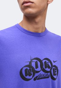 Κοντινό πλάνο ενός ατόμου που φοράει μοβ μπλουζάκι Nike με μαύρο κυκλικό γράμμα που γράφει "Nike Athletics" στο στήθος.