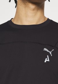 Svart atletisk tröja med ribbat mönster, har rundad hals, kontrasterande sömmar och silverfärgade logotypdetaljer på bröstet.