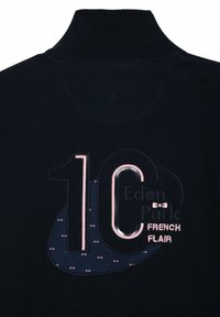 Pull bleu marine avec un col haut. Il présente un "10" et "Eden Park" brodés en rose clair, entourés d'un accent circulaire bleu marine.