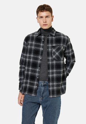 Jeune homme portant une chemise à carreaux noire et blanche à boutons par-dessus un col roulé gris foncé et un jean bleu, debout sur un fond uni.