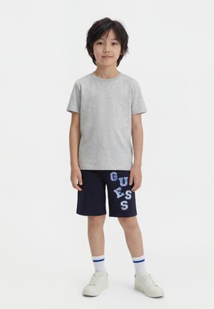 Niño de pie mirando hacia adelante, vistiendo una camiseta gris, pantalones cortos azul marino con letras "GUESS", calcetines blancos con rayas azules y zapatillas blancas.