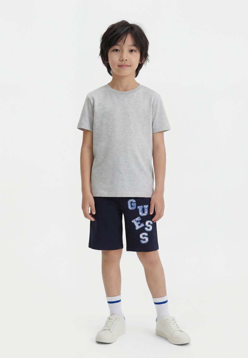 Garçon debout, face à l’avant, portant un t-shirt gris, un short bleu marine avec l’inscription « GUESS », des chaussettes blanches avec des rayures bleues et des baskets blanches.