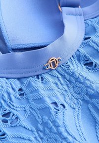 Hellblauer Stoff mit komplizierter Stickerei und ausgeschnittenen Mustern, akzentuiert durch einen glatten, breiteren Riemen und ein goldfarbenes Verschlussdetail.