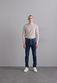 Mann, der gegen eine schlichte graue Wand steht und einen hellbeigen Rollkragenpullover, dunkelblaue Jeans und weiße Sneakers trägt. Er blickt nach vorne, mit den Händen an den Seiten.