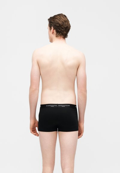 Άνδρας με κοντά καστανά μαλλιά, που φαίνεται από πίσω, φοράει μαύρα μπόξερ με τύπωμα στη μέση, σε μονόχρωμο φόντο.