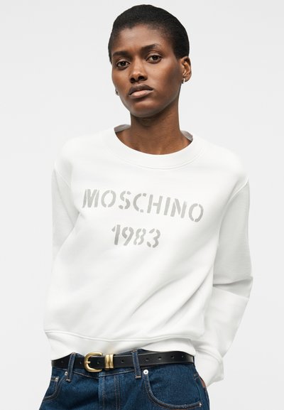 Νεαρή γυναίκα που φοράει λευκό φούτερ Moschino 1983, μπλε τζιν και μαύρη ζώνη με χρυσή αγκράφα σε απλό φόντο.