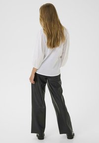 Frau mit langem, hellbraunem Haar, die eine weiße Bluse mit Puffärmeln und schwarze, weit geschnittene Lederhosen trägt, steht vor einem schlichten Hintergrund.