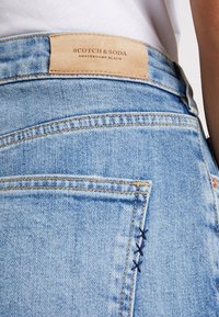 Jeans de mezclilla de color azul claro con una etiqueta de cuero marrón en la cintura, que presentan costuras en contraste y un bordado decorativo en azul marino.