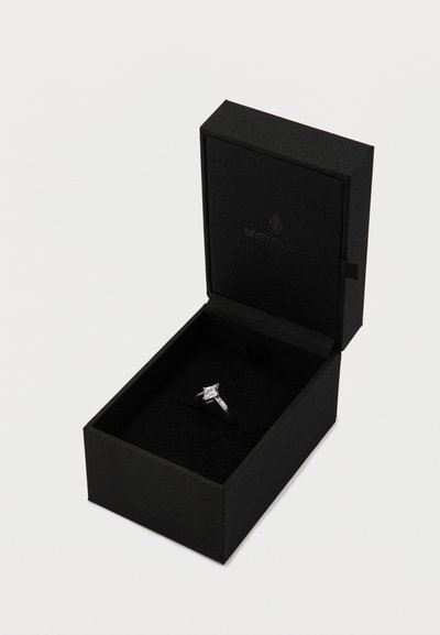 Fekete gyűrűdoboz, puha belsővel, egy gyémántgyűrűnek ad otthont, amelynek központi köve négyzet alakúra van csiszolva, két oldalsó kővel együtt, mindez ezüst foglalatban.