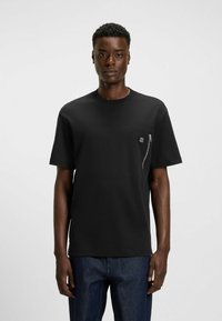 T-shirt nera a maniche corte con scollatura rotonda, dotata di tasca con zipper sul petto e un accento logo testurizzato. Tessuto liscio, vestibilità comoda.