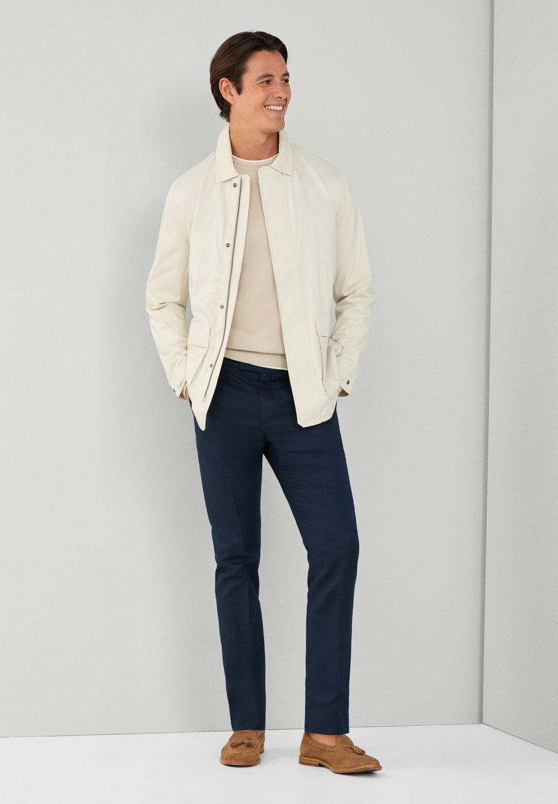 Veste beige clair avec un col, portée sur un pull crème, associée à un pantalon bleu marine foncé. Des mocassins bruns complètent la tenue.