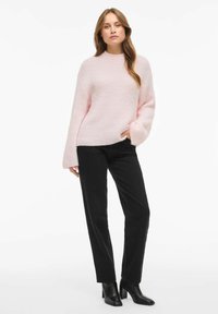 Jasnoróżowy sweter o prążkowanej fakturze i długich rękawach, zestawiony z czarnymi jeansami o prostych nogawkach oraz czarnymi botkami na obcasie. Gładkie tło.
