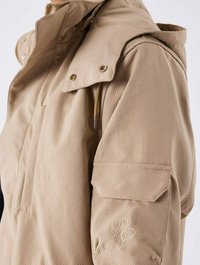 Beige jakke med struktureret stof, trykknapper og lynlåslukning foran. Inkluderer et broderet logo på ærmelommen.