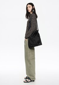 Czarna torba typu tote z podwójnymi uchwytami, teksturowany materiał i logo. Modelka nosi wzorzysty sweter, zielone spodnie w szerokim kroju i ozdobne sandały.