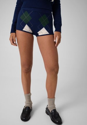 Personne portant un short en maille bleu marine avec un motif losanges vert et blanc, des chaussettes beiges et des mocassins noirs sur fond gris.