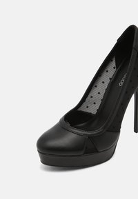 Escarpin à plateforme noir à talon haut avec bout arrondi et côtés en mesh transparent ornés de petits pois noirs.
