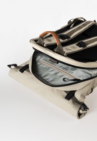 Beige Rucksack mit schwarzem Innenraum, ausgestattet mit einem Handgriff oben, verstellbaren Trägern und einer silbernen Reißverschlusstasche im Inneren. Glattes, langlebiges Material.