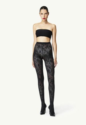 Frau steht, trägt ein schwarzes Bandeau-Top, schwarze, durchsichtige Strumpfhose mit Blumenmuster und schwarze High Heels vor weißem Hintergrund.