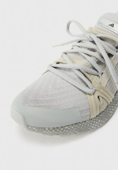 Light grey sneaker featuring a textured fabric upper, elastic straps, and a soft rubber toe cap, with a speckled midsole design.