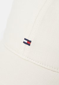 Gorra beige de tela texturizada, con un pequeño logo bordado que presenta franjas rojas, blancas y azules en el lado izquierdo frontal.