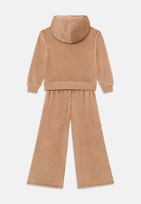Survêtement en velours beige comprenant un haut à capuche et un pantalon à jambes larges. Texture douce avec des poignets et un ourlet côtelés, offrant une coupe décontractée.