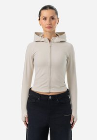 Beige skrátená mikina s kapucňou a dlhými rukávmi, vyrobená z elastického materiálu s otvormi na palce, skombinovaná s tmavými džínsami.