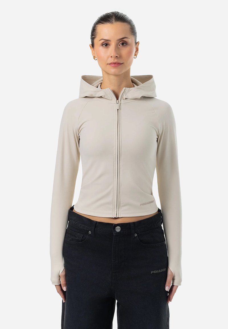 Beige skrátená mikina s kapucňou a dlhými rukávmi, vyrobená z elastického materiálu s otvormi na palce, skombinovaná s tmavými džínsami.