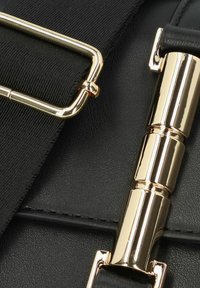 Dettaglio di una borsa a tracolla in pelle nera con un fermaglio in metallo dorato e una tracolla testurizzata. La superficie è liscia con cuciture visibili.