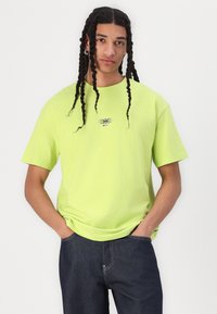 Neon green cotton t-shirt with a round neck, short sleeves, and a small graphic print at the center. Styled with dark denim jeans.
