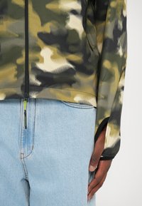 Camouflage jacket in shades of green and beige with a zip front, long sleeves, and side slits, paired with light blue jeans.