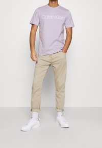 Camiseta de algodón lavanda con logo "Calvin Klein", combinada con chinos beige claro y zapatillas blancas, con puños enrollados y calcetines blancos.