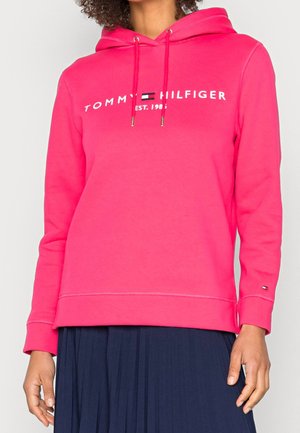 Kvinde iført en knaldpink Tommy Hilfiger hættetrøje med logo og mørkeblå plisseret nederdel, stående i afslappet kropsholdning.