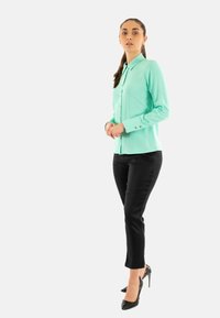 Donna in piedi con camicia verde menta a bottoni, pantaloni neri aderenti e tacchi alti neri, su uno sfondo chiaro e semplice.