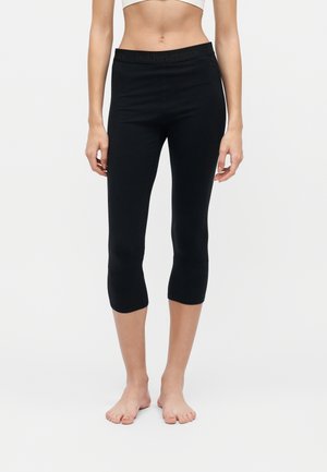 Femme portant des leggings noirs courts et un top blanc court, debout pieds nus sur un fond uni.