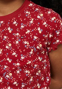 Rotes geripptes T-Shirt mit kurzen rüschigen Ärmeln, das ein florales Muster in Gelb, Weiß und Blau zeigt, sowie einem kleinen Logo-Detail in der Mitte.