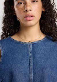Young person with curly dark hair and freckles wearing blue denim top and thin gold necklace against white background.