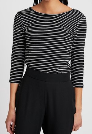 Personne portant un haut à manches longues rayé horizontalement noir et blanc et un pantalon noir taille haute, les mains détendues le long du corps.