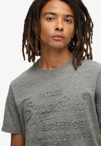 Grå bomulds t-shirt med prægede logo-tekster. Korte ærmer med klassisk rund hals, glat tekstur og afslappet pasform.