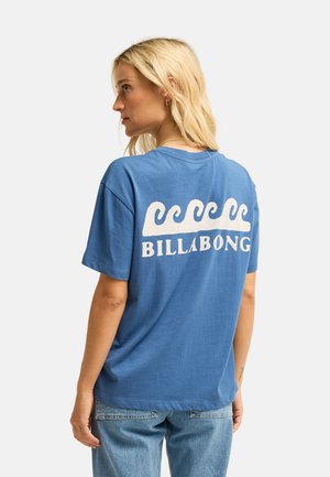 Blondynka w niebieskim t-shircie Billabong z białymi wzorami fal i tekstem na plecach, dobrana do jasnoniebieskich dżinsów.