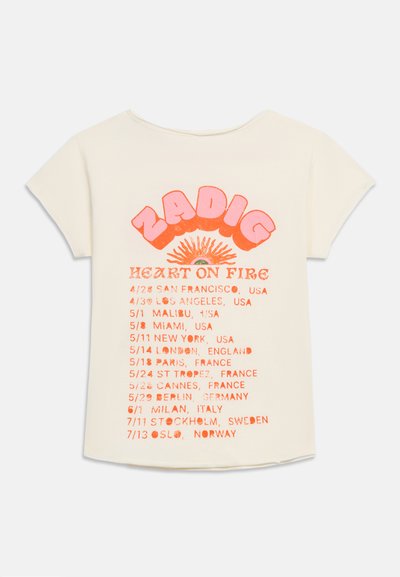 Κρέμ μπλουζάκι με ροζ κείμενο "ZADIG", πορτοκαλί γραφικό ήλιου και λίστα ημερομηνιών περιοδείας με μεγάλες πόλεις των ΗΠΑ και της Ευρώπης στην πίσω πλευρά.