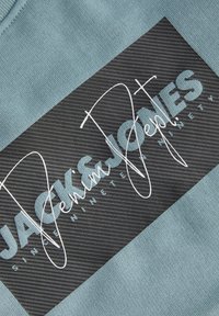 Ύφασμα φούτερ ανοιχτού μπλε με υφή, με μαύρη πινακίδα που φέρει τη φράση «JACK & JONES» με έντονα λευκά γράμματα και σχέδιο σε στιλ υπογραφής.