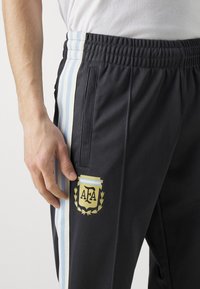 Pantalon de sport noir avec des rayures latérales bleu clair et blanches. Présente un emblème jaune avec "AFA" et un design décoratif de laurier.