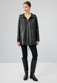 Giubbotto in pelle nera con cappuccio, chiusura con zip, due tasche frontali e orlo regolabile. Indossato sopra una maglietta leggera e abbinato a leggings neri e stivali.