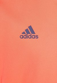 Camiseta deportiva color coral hecha de tela texturizada con el logo de Adidas en azul. El diseño incluye tres franjas diagonales por encima del logo.