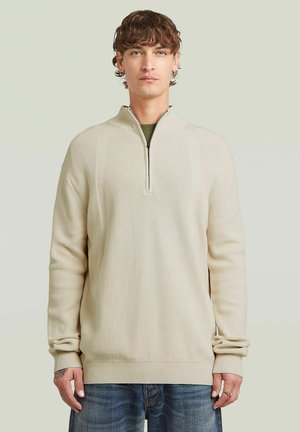 Jonge man met krullend haar die een beige sweater met kwartrits en blauwe spijkerbroek draagt, tegen een effen lichte achtergrond.