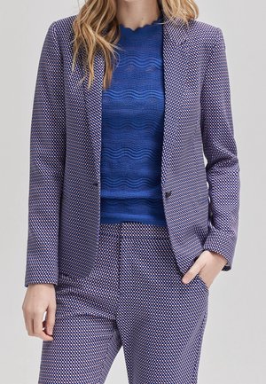 Femme portant un haut bleu texturé et un blazer ainsi qu'un pantalon assortis à motifs violets, la main dans la poche, avec des cheveux blonds ondulés.