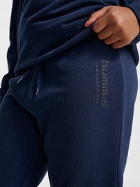 Enfant portant un pantalon de sport bleu marine avec la marque "hummel SPORTSWEAR", remontant un sweat-shirt bleu marine assorti.