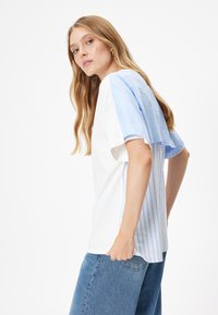 Hvid T-shirt med blå stribede ærmer og sideløsninger. Har en afslappet pasform og blød stofstruktur. Simpel, moderne design.