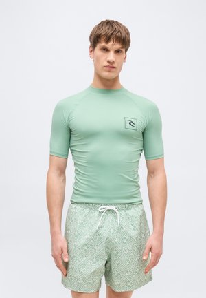 Fiatal férfi világos zöld, testhezálló rash gard viselésével és mintás fürdőnadrágban, melynél húzózsinór is van, egyszerű háttér előtt áll.